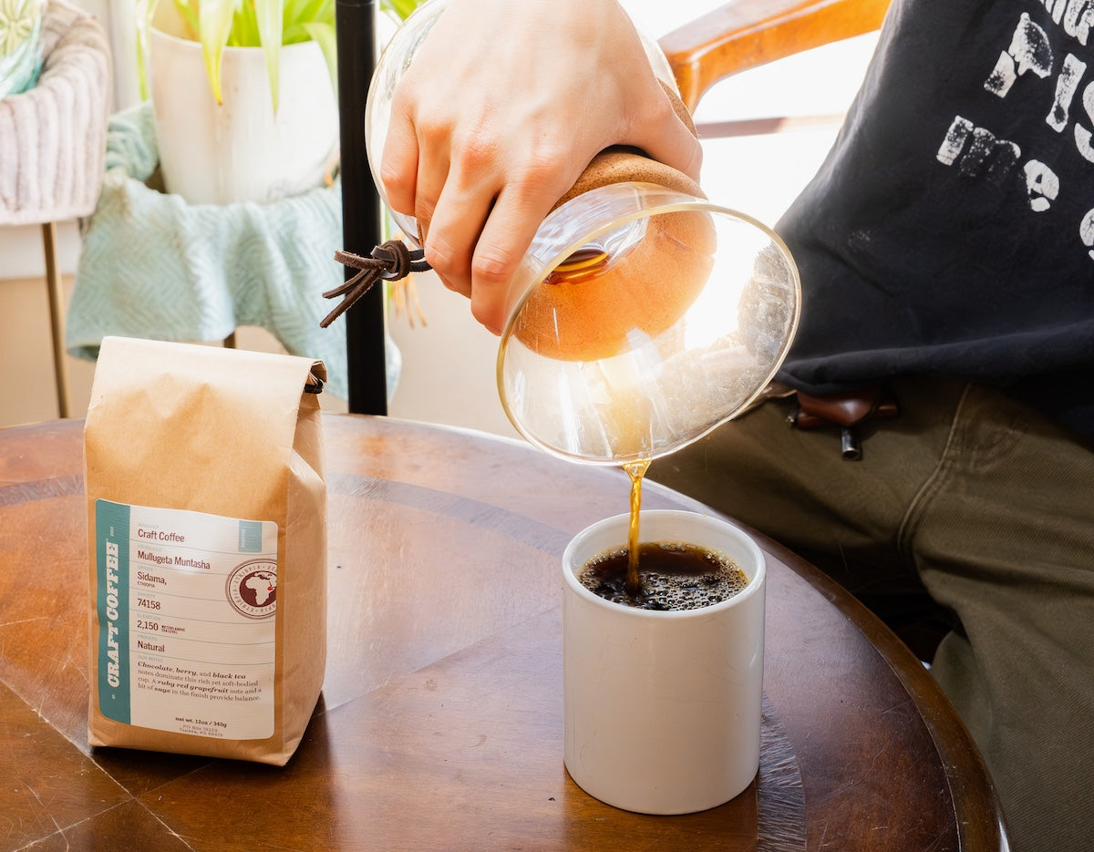 Person pouring coffee from a glass Chemex carafe into a white cup on a wooden table with a coffee bag nearby.