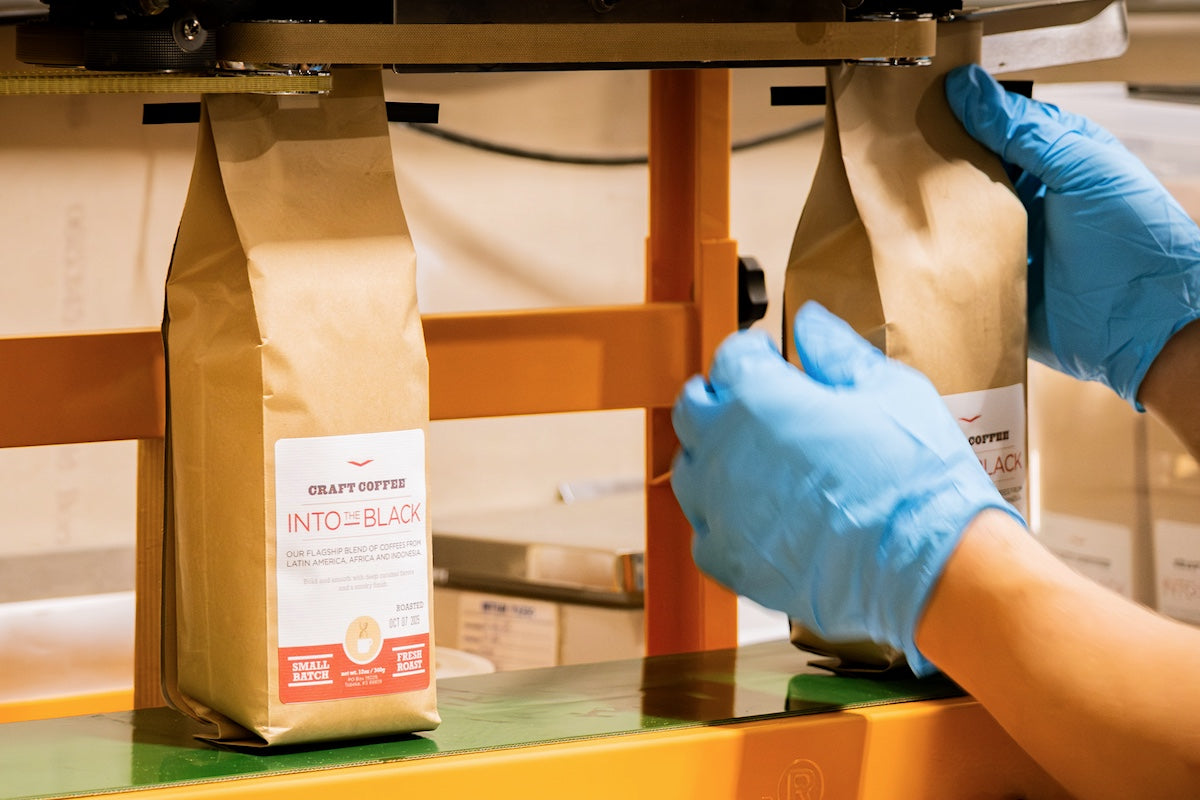 Person wearing blue gloves sealing coffee bags labeled 'Into the Black' on a conveyor belt.