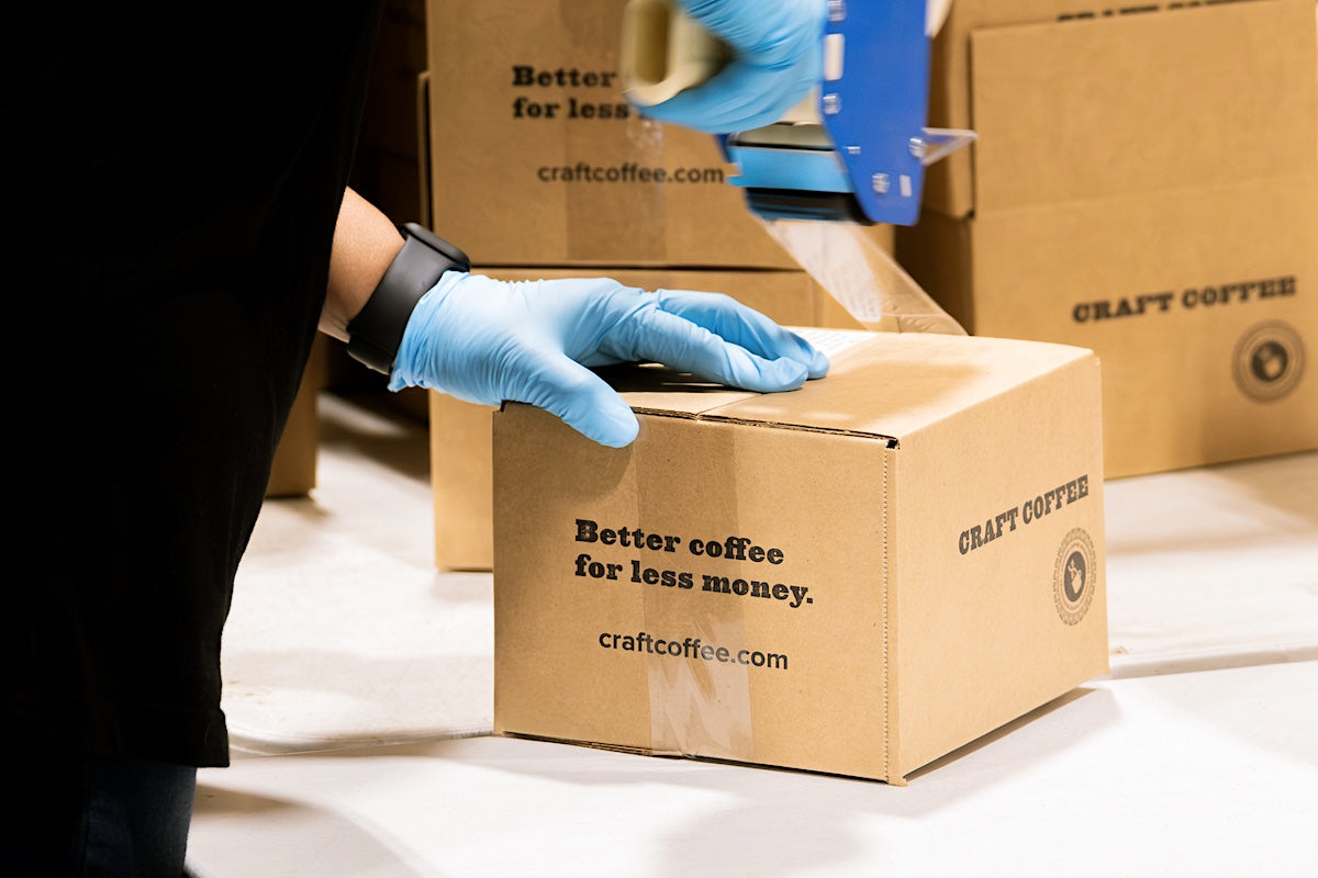 Person wearing gloves packaging a cardboard box with 'Craft Coffee' branding.